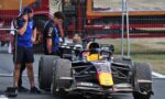 Formula 1 car with yellow nose parked behind a barrier, crew standing nearby in blue team uniforms, one on a phone.