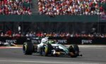 Formula 1 car speeding along the track with a packed grandstand crowd in the background near the pit wall nearby the stands.
