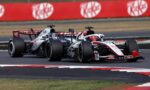 Two Formula 1 cars race side by side on a track, with a red barrier wall in the background and green grass beyond it.