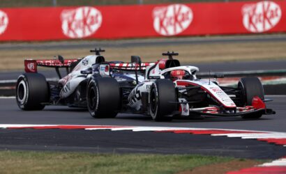 Two Formula 1 cars race side by side on a track, with a red barrier wall in the background and green grass beyond it.