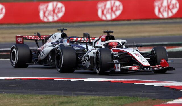 Two Formula 1 cars race side by side on a track, with a red barrier wall in the background and green grass beyond it.