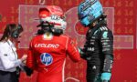 Two Formula 1 drivers in helmeted racing suits face each other, red Ferrari driver on the left and a black-teal suit on the right, against a red numbered backdrop, with a staff member on the left.