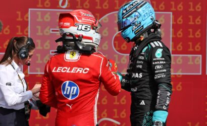 Two Formula 1 drivers in helmeted racing suits face each other, red Ferrari driver on the left and a black-teal suit on the right, against a red numbered backdrop, with a staff member on the left.