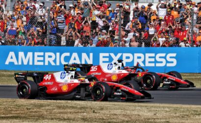 Two red Ferrari Formula 1 cars race side by side on track as a cheering crowd watches behind a chain-link fence and a blue barrier reading American Express.