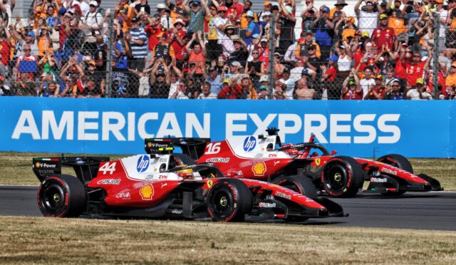 Two red Ferrari Formula 1 cars race side by side on track as a cheering crowd watches behind a chain-link fence and a blue barrier reading American Express.
