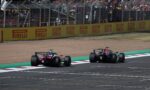Two Formula 1 cars race on a track beside a yellow Pirelli barrier with a crowded grandstands in the background.