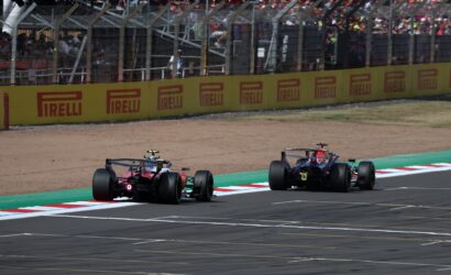 Two Formula 1 cars race on a track beside a yellow Pirelli barrier with a crowded grandstands in the background.