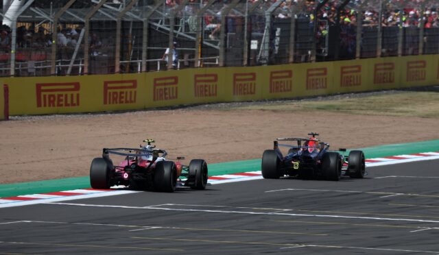 Two Formula 1 cars race on a track beside a yellow Pirelli barrier with a crowded grandstands in the background.