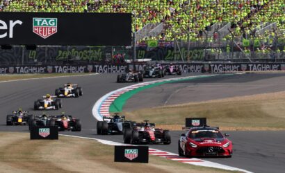 Motor racing scene with a packed grandstand of fans in bright yellow-green shirts; Formula 1 cars on a curving track and a TAG Heuer advertising barrier.