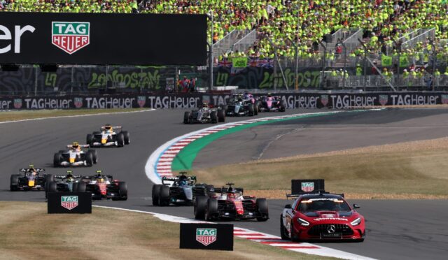 Motor racing scene with a packed grandstand of fans in bright yellow-green shirts; Formula 1 cars on a curving track and a TAG Heuer advertising barrier.