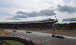 Formula 1 cars race on a track near a grandstand under a cloudy sky with spectators in the stands.