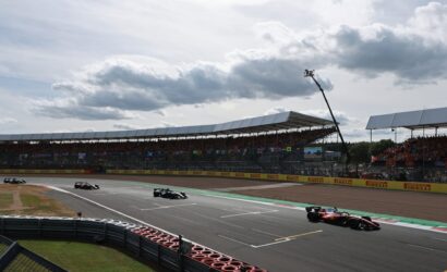 Formula 1 cars race on a track near a grandstand under a cloudy sky with spectators in the stands.