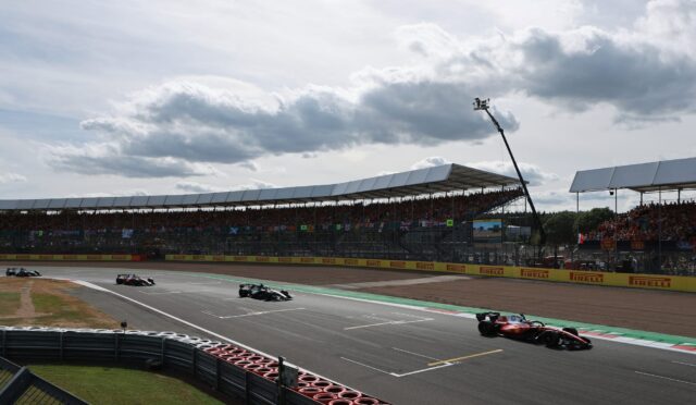 Formula 1 cars race on a track near a grandstand under a cloudy sky with spectators in the stands.