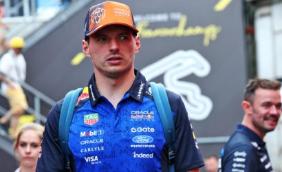 Male racer in blue Oracle Red Bull Racing shirt with sponsor logos, orange cap, and blue backpack straps at a racing event, looking to the side.