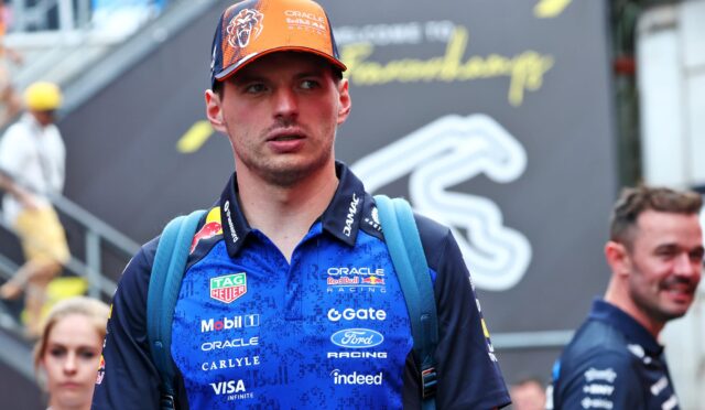 Male racer in blue Oracle Red Bull Racing shirt with sponsor logos, orange cap, and blue backpack straps at a racing event, looking to the side.