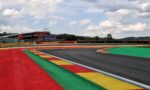 Race track with red, green, yellow, and blue run-off areas and a grandstand in the distance under a cloudy sky
