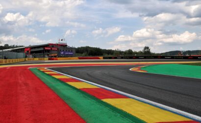 Race track with red, green, yellow, and blue run-off areas and a grandstand in the distance under a cloudy sky