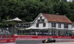 Single-seater race car on a track past red Moët & Chandon barriers with spectators and a white half-timbered building in the background.