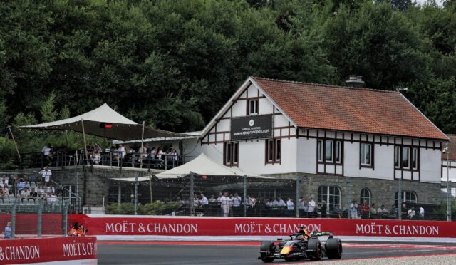 Single-seater race car on a track past red Moët & Chandon barriers with spectators and a white half-timbered building in the background.