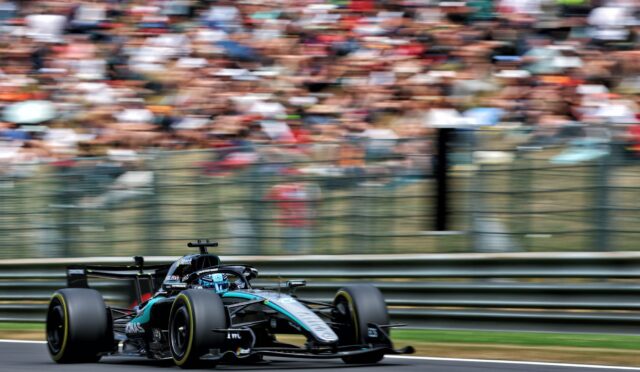 Formula 1 race car (Mercedes) speeding along track with blurred grandstands in the background, capturing high-speed motion.