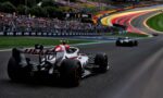 Formula 1 race car on a curving track with a packed grandstand in the background and colorful barrier walls nearby.