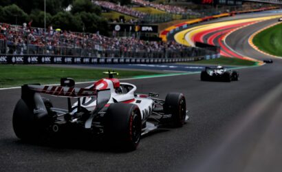 Formula 1 race car on a curving track with a packed grandstand in the background and colorful barrier walls nearby.