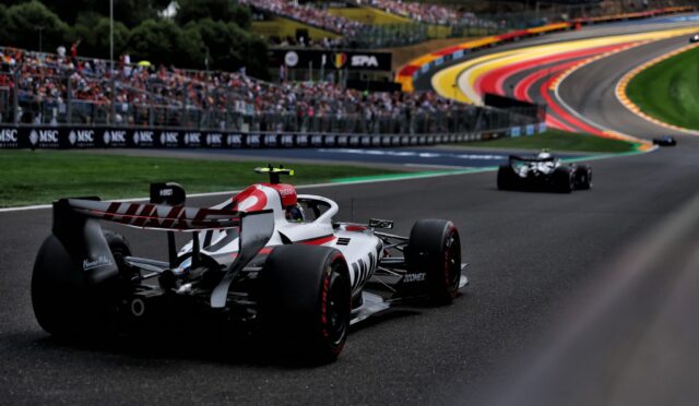 Formula 1 race car on a curving track with a packed grandstand in the background and colorful barrier walls nearby.