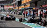 Formula 1 cars in the pit lane with teams and crew nearby, ready for a stint on the track, banners overhead and garages in the background.