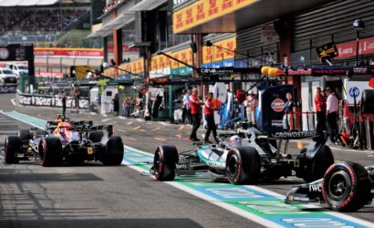 Formula 1 cars in the pit lane with teams and crew nearby, ready for a stint on the track, banners overhead and garages in the background.