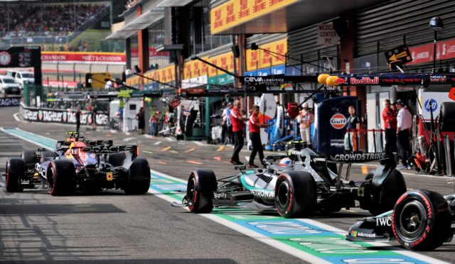 Formula 1 cars in the pit lane with teams and crew nearby, ready for a stint on the track, banners overhead and garages in the background.