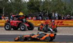 Red tow truck with a deployed crane on a race track, orange-clad marshals and a damaged car being moved while spectators watch behind a safety fence.
