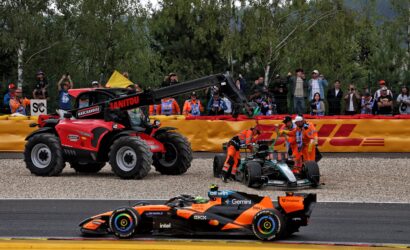 Orange Formula 1 car speeding on track with a red tow crane nearby and spectators behind a fence barrier.