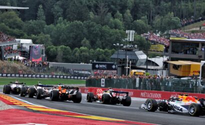 Formula 1 cars accelerating off the start on a bright track, with grandstands full of spectators and forest backdrop in the background