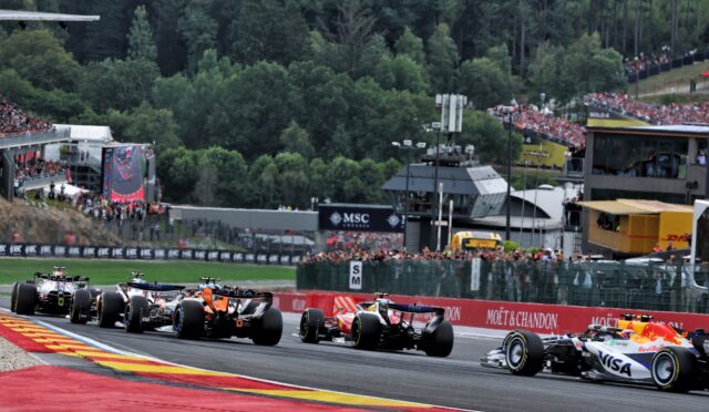 Formula 1 cars accelerating off the start on a bright track, with grandstands full of spectators and forest backdrop in the background