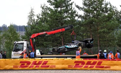 Crane lifts a race car onto a truck behind a DHL barrier, with pit crew wearing orange vests nearby in a wooded area.