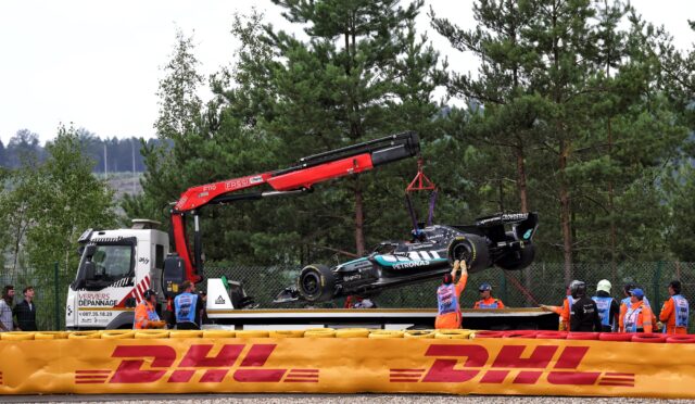 Crane lifts a race car onto a truck behind a DHL barrier, with pit crew wearing orange vests nearby in a wooded area.