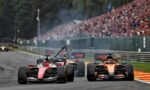 Front-line Formula 1 cars race down a track with a packed grandstand of spectators in the background.