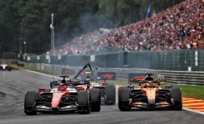 Front-line Formula 1 cars race down a track with a packed grandstand of spectators in the background.