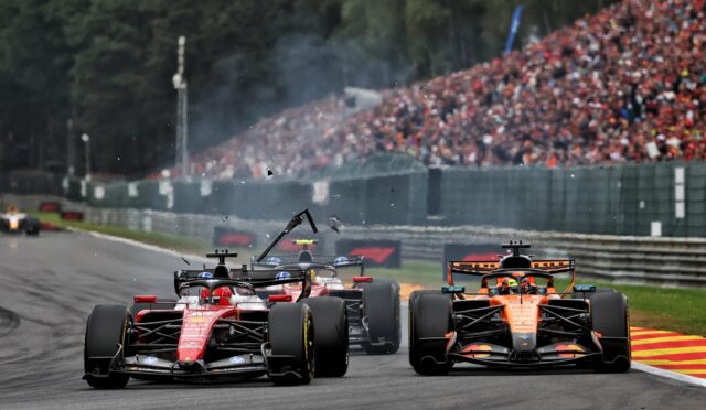 Front-line Formula 1 cars race down a track with a packed grandstand of spectators in the background.