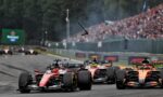 Formula 1 cars race down the straight with a packed grandstand in the background; debris or parts soar through the air above the track.