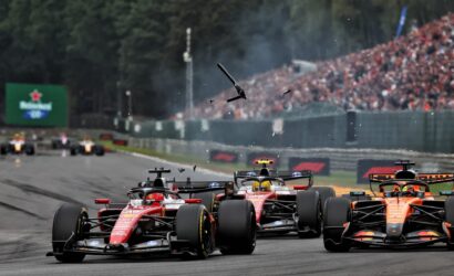 Formula 1 cars race down the straight with a packed grandstand in the background; debris or parts soar through the air above the track.
