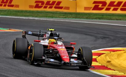Red Formula 1 car rounding a bend on a race track, with yellow-red curbs and DHL banners in the background.