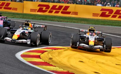 Formula 1 race cars cornering through a chicane with DHL barriers and a cheering crowd in the background.