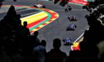Race cars navigate a tight curve on a race track, with vibrant red, yellow, and green curbs and silhouetted spectators in the foreground.