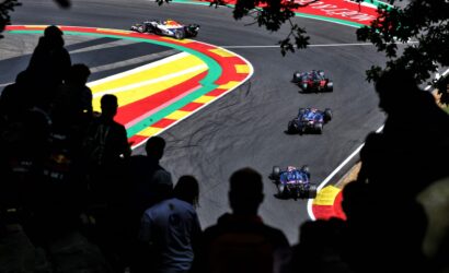 Race cars navigate a tight curve on a race track, with vibrant red, yellow, and green curbs and silhouetted spectators in the foreground.