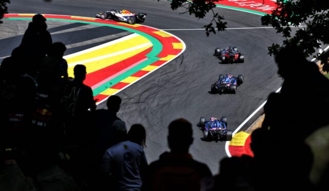 Race cars navigate a tight curve on a race track, with vibrant red, yellow, and green curbs and silhouetted spectators in the foreground.