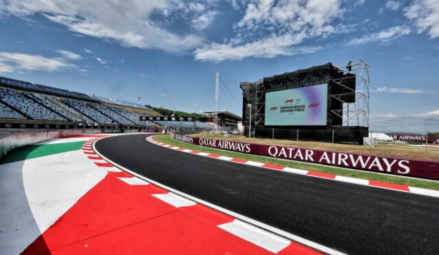 Race track with red-and-white curbs, empty grandstand, and a large stage screen displaying event graphics.