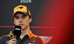 Formula 1 driver in an orange cap and matching team jacket speaks into a microphone at a press event, with '2026' on a dark backdrop behind.
