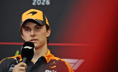 Formula 1 driver in an orange cap and matching team jacket speaks into a microphone at a press event, with '2026' on a dark backdrop behind.