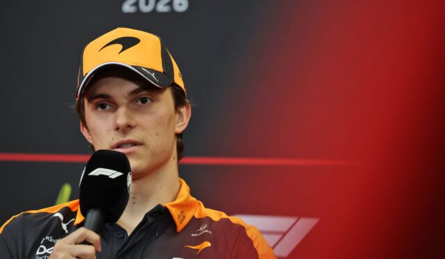 Formula 1 driver in an orange cap and matching team jacket speaks into a microphone at a press event, with '2026' on a dark backdrop behind.
